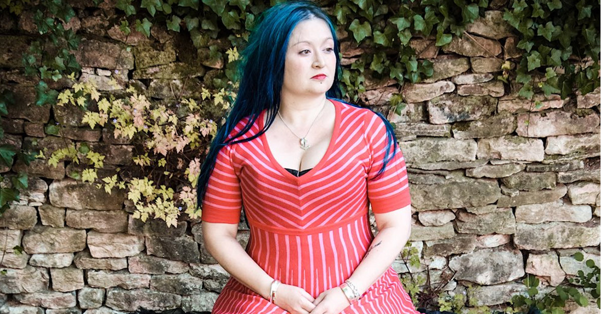 Eliza Carthy - wearing red dress with drystone wall behind